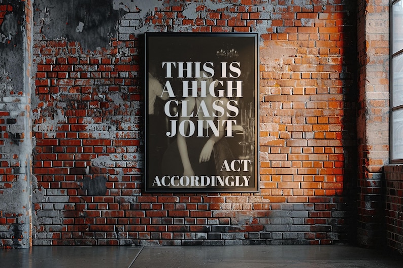 May include: A framed black and white poster with the text "THIS IS A HIGH CLASS JOINT ACT ACCORDINGLY" hangs on a brick wall. The brick wall is red and brown, with a concrete floor. A window is visible on the right.