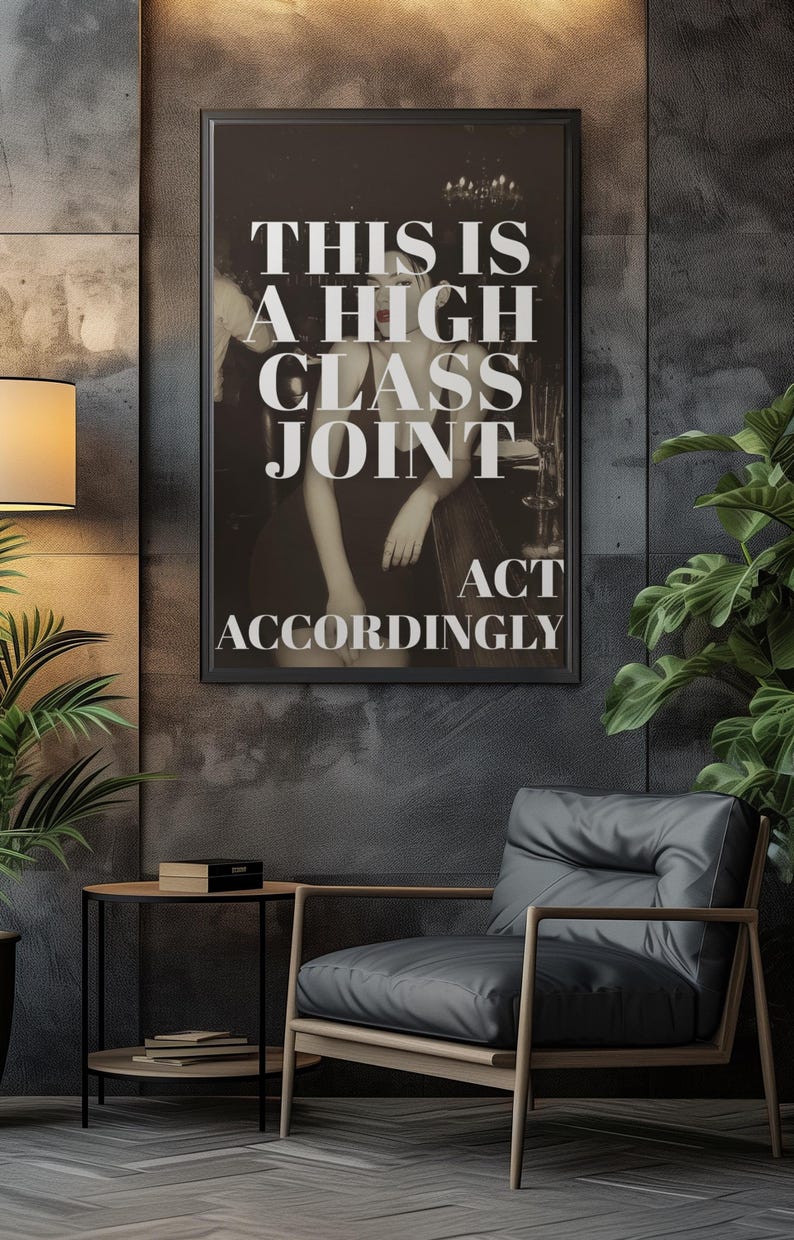 May include: A framed black and white print with the text "THIS IS A HIGH CLASS JOINT ACT ACCORDINGLY." The print hangs on a dark gray textured wall. A modern chair and side table are in the foreground.