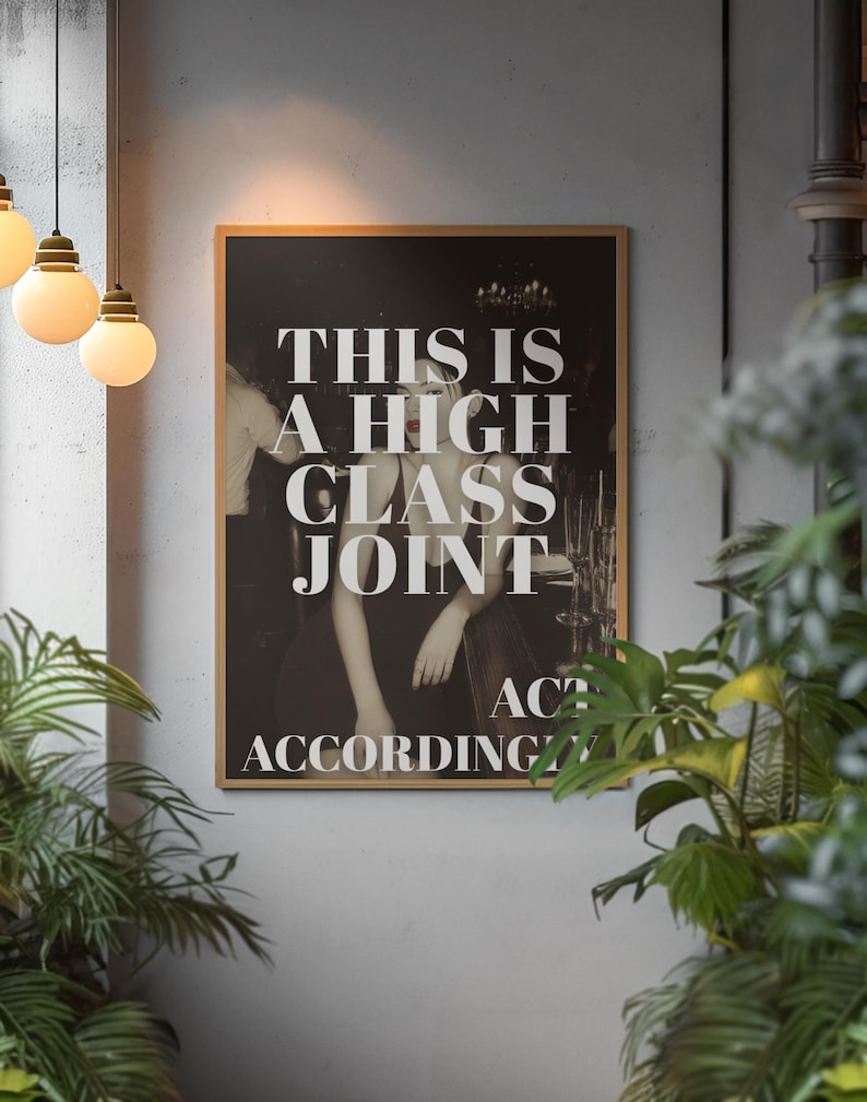 May include: A framed black and white print with the text "THIS IS A HIGH CLASS JOINT ACT ACCORDINGLY". The image features a person in a dress leaning on a bar. The print is in a room with hanging lights and plants.
