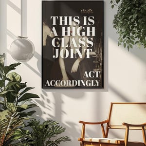 May include: Black framed poster with the text "THIS IS A HIGH CLASS JOINT ACT ACCORDINGLY" in white. The poster is hanging on a white wall, with a wooden chair and plants in the foreground. The image has a vintage aesthetic.