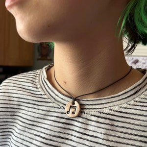 May include: A black cord necklace with a round, beige pendant featuring a black musical note design.