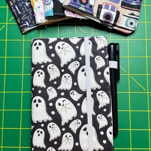 May include: A black and white notebook cover with a pattern of white ghosts on a black background. The cover has a white elastic band closure and a black pen is tucked into the elastic band.