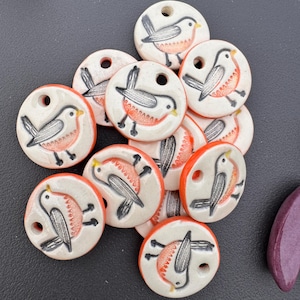 May include: A collection of ceramic beads with a bird design. Each round bead features a white base with a black and orange bird illustration and an orange border. The beads have a hole for stringing and are suitable for jewelry making or crafts.