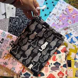 May include: A black drawstring pouch featuring a repeating white cat pattern with various textures, held by a white cord. The pouch is surrounded by multiple folded fabric pieces displaying diverse cat-themed prints in colors like pink, yellow, and blue.