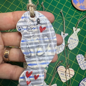 May include: A ceramic ornament shaped like a manatee with blue and white stripes and red hearts. The ornament is hanging from a twine string. The ornament is labeled "Welaka Cat Lady".