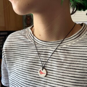 May include: A round, white pendant necklace with a black cord. The pendant features a red cardinal bird design. The person wearing the necklace is wearing a black and white striped shirt.