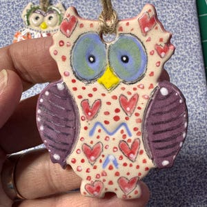 May include: A ceramic owl ornament with a jute string for hanging. The owl features a cream-colored body with red hearts and dots, purple wings, blue eyes, and a yellow beak. A second owl ornament is visible in the background.