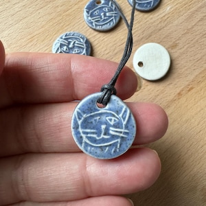 May include: A blue ceramic necklace with a cat face pendant. The pendant is round and has a white glaze with a blue cat face design. The necklace is on a black cord.