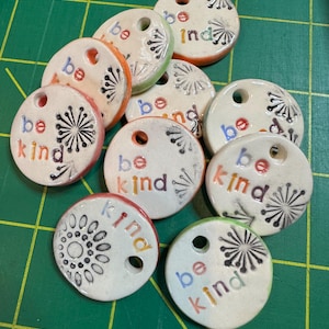 May include: Nine white ceramic round tags with holes for string. Each tag has a black and white dandelion design and the words "be kind" in colorful letters.