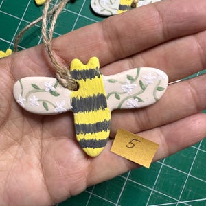 May include: A bee-shaped ornament made of off-white material with a yellow and black striped body. The wings feature a painted green vine pattern with small white flowers. A jute twine loop is attached to the top, and a small yellow tag with the number 5 is visible.