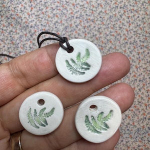 May include: Three white ceramic pendants with green fern designs. Two pendants have a hole for a cord, and one pendant has a cord attached.