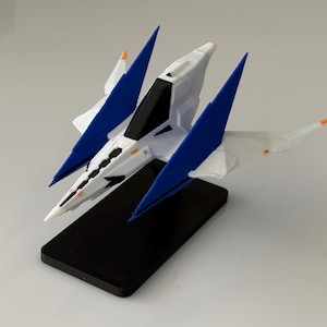 May include: A white and blue model spaceship featuring a pointed nose, angular wings, and a black cockpit canopy. The spacecraft is mounted on a rectangular black base, showcasing a sleek design with small orange accents on the wingtips.