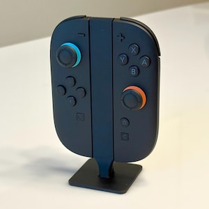 May include: A dark blue game controller with a black base. The controller has a blue joystick on the left and an orange joystick on the right. It features buttons in white, blue, and orange.