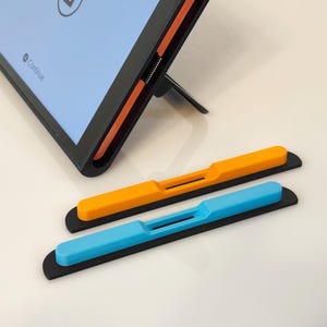 Op de afbeelding: Een tablet met een zwart frame en oranje accenten wordt ondersteund door een standaard. Twee tabletaccessoires, één oranje en één blauw, zijn eronder geplaatst. De accessoires hebben een zwarte basis en een rechthoekig ontwerp.