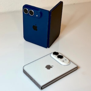 Può includere: Uno smartphone pieghevole blu con un modulo fotocamera a doppia lente e un logo Apple nero, posizionato in verticale, accanto a uno smartphone pieghevole bianco con un modulo fotocamera simile e un logo Apple argento, appoggiato in piano.