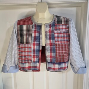 May include: A cropped jacket featuring a patchwork design of various red, white, blue, and black plaid patterns. The garment has three-quarter length sleeves with light blue cuffs, a collarless round neckline, and a quilted texture throughout the body.