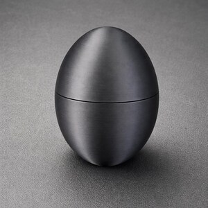May include: A dark grey, egg-shaped container with a brushed metal finish. The container is split in the middle, suggesting it opens. It sits on a textured grey surface.