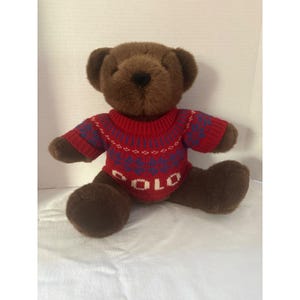 May include: A brown plush teddy bear wearing a red knit sweater with a blue and white geometric pattern. The sweater features the word POLO in white capital letters across the chest. The bear has dark brown fur, rounded ears, and a black nose.