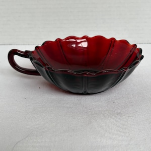 May include: A ruby red glass bowl with a handle. The bowl has a scalloped edge and a petal-like design. The handle is curved and attached to the side of the bowl. The bowl is sitting on a white surface.