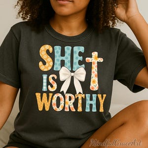 May include: Dark grey t-shirt with the words "SHE IS WORTHY" in a colourful, floral design. The text features a white bow and various patterns, including flowers and gingham. The shirt is a casual wear item.