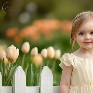 May include: A young girl with blonde hair and a white flower in her hair is wearing a pale yellow dress. She is standing in front of a white picket fence with a field of tulips behind her.