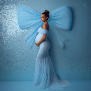Può includere: Un abito premaman azzurro con design a spalle scoperte e gonna a sirena fluida. L'abito è adornato con abbellimenti scintillanti. Un grande fiocco azzurro è montato sulla parete dietro il soggetto, creando un'estetica morbida e sognante.