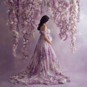 May include: A pregnant person wearing a flowing, off-the-shoulder gown with a floral pattern in shades of purple and white. The gown's train trails behind. The background features hanging purple and white flowers.