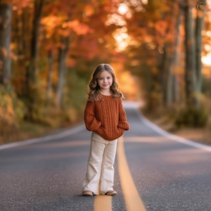 May include: A child wearing a long-sleeved, cable-knit jumper in a burnt orange colour paired with cream-coloured, wide-leg trousers. The jumper features a textured vertical cable pattern and a crew neckline, while the trousers have a clean, straight-leg fit.
