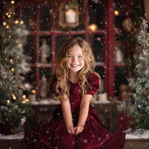 Peut inclure: Une jeune fille aux longs cheveux blonds ondulés sourit, vêtue d'une robe de velours bordeaux. L'arrière-plan présente des décorations de Noël, dont de petits arbres à feuilles persistantes et une fenêtre encadrée de rouge, avec de la neige qui tombe.