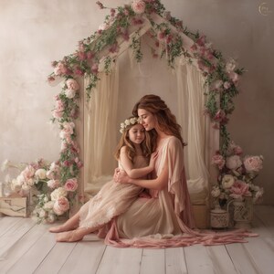 May include: A mother and child embrace in a soft, romantic setting. The scene is framed by a white structure adorned with pink and white flowers. The mother wears a flowing pink dress, and the child wears a matching dress and flower crown. The background is a neutral color.