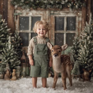 Puede incluir: Un niño con una camisa beige y un peto de pana verde posa junto a un cervatillo en un escenario nevado. El fondo presenta árboles de Navidad y una puerta de madera. El niño sonríe, creando una escena festiva.