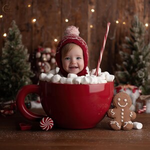 May include: A smiling baby wearing a red knit hat sits in a giant red mug filled with marshmallows. A red and white striped straw and a gingerbread man are next to the mug. Christmas-themed photo.