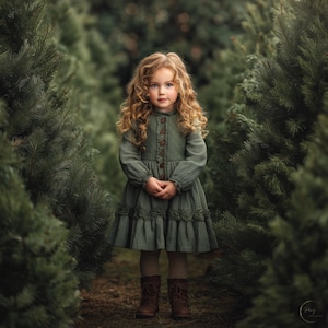 Puede incluir: Una niña con largo cabello rubio rizado está de pie en un bosque de árboles verdes. Lleva un vestido verde de pana con botones y botas marrones. El vestido tiene un dobladillo con volantes y mangas largas. La niña mira directamente a la cámara.
