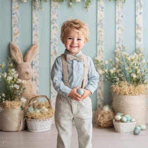 May include: A young child wearing a light blue striped shirt, braces, and a bow tie, holding a blue egg. The child smiles in front of an Easter backdrop with a bunny, baskets, and eggs.