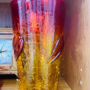 Pode incluir: Um vaso cilíndrico de vidro com textura craquelada e um gradiente de cor que transita do vermelho profundo no topo para um amarelo âmbar quente na base. Dois elementos decorativos de vidro em forma de folha estão aplicados nas laterais.