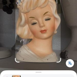 Könnte beinhalten: Eine Vintage-Kopfvase aus Keramik mit einer Frau mit geschlossenen Augen, blonden Haaren und einem Blumenkranz. Das Gesicht ist mit zarten Zügen und rosigen Wangen bemalt. Ein dekorativer Artikel.