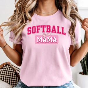 Pode incluir: T-shirt rosa claro com as palavras "SOFTBALL MAMA" em negrito, rosa, com uma fonte em estilo universitário. A palavra "MAMA" está numa caixa rosa com um laço. A t-shirt é de cor rosa claro.