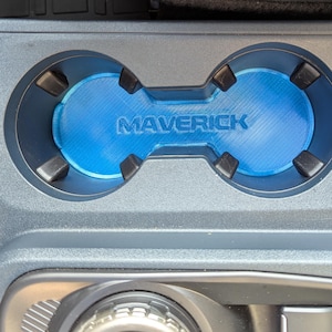 May include: A custom-fit car cup holder insert with the word "MAVERICK" in raised lettering. The insert is blue and fits into a gray car console. The cup holder has two circular openings.