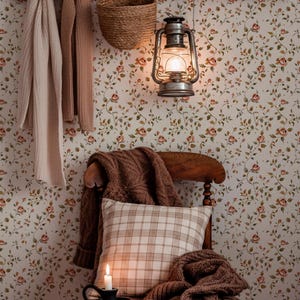 May include: A cozy interior scene featuring a wooden chair with a plaid pillow and a brown knitted blanket. A lit candle sits on a small black dish. A vintage lantern hangs from the wall, and a floral wallpaper provides a warm backdrop. Scarves and a basket hang on a wooden rack.