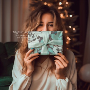 May include: A woman is holding a blue and gray gift box with a white ribbon in front of her face. The gift box has a floral pattern.