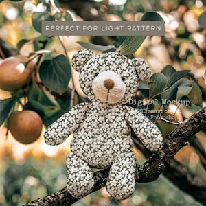May include: A grey teddy bear with a white floral pattern sits on a tree branch. The bear has a brown nose and brown eyes. The background is a blurry image of green leaves and apples.