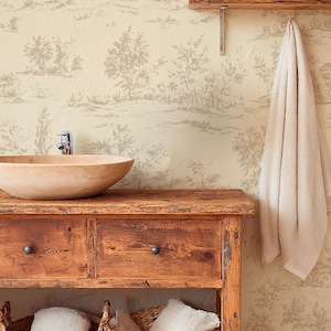 Puede incluir: Una escena de baño rústica con un tocador de madera con un lavabo sobre encimera, dos cajones y dos cestas de mimbre. El papel pintado tiene un diseño toile de Jouy. Una estantería contiene toallas enrolladas y una toalla cuelga de la pared.