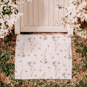 May include: A white doormat with a floral pattern in shades of brown and blue. The doormat is placed on a patch of green grass.