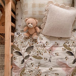 May include: A beige teddy bear rests on a bed with a floral quilt and a cream crocheted pillow. The quilt displays pink birds and white flowers. The pillow has a scalloped edge. The bed frame is made of light-coloured wood.