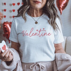 May include: White t-shirt with the word "Valentine" written in pink cursive with two small pink hearts above the word.