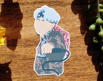 May include: A sticker featuring a stylized illustration of a person with light blue hair and a bandana. The person is holding a microphone. The design incorporates patterns in shades of blue, red, and gray. The sticker is on a wooden surface.