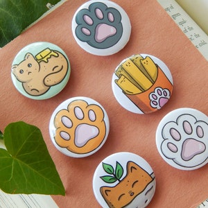 May include: Set of six 1-inch button pins featuring cute cat illustrations. The designs include a cat with butter, a cat with a plant, a cat with fries, and three paw prints in different colors.