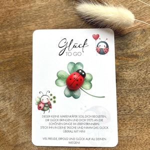 Op de afbeelding: Een kleine rode lieveheersbeestje-hanger met zwarte stippen op een groen vierbladig klavertje vier. De hanger is bevestigd aan een witte kaart met de tekst "Glück TO GO" en een boodschap in het Duits over geluk.
