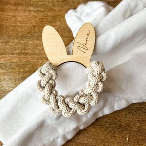 Könnte beinhalten: Ein Napfkinring aus Holz in Form eines Hasen mit dem eingravierten Namen "Nina". Der Ring hält eine weiße Serviette und liegt auf einer Holzoberfläche.