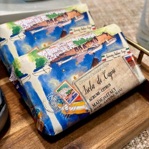 Può includere: Due saponette italiane, ciascuna avvolta in carta con un'illustrazione di Capri. La confezione presenta il testo "Isola di Capri" e "Luxury Italian Soap". Le saponette sono esposte su un vassoio di legno.